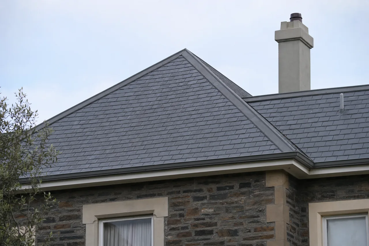 A hipped slate roof on a bluestone villa with a rendered chimney against a pale sky — slate roofing, Adelaide. Illustrative example.